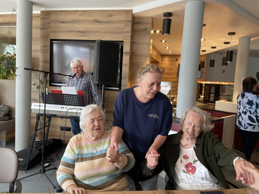 Drei ältere Frauen, zwei sitzend und eine stehend, lächeln und halten sich an den Händen in einer fröhlichen Innenumgebung. Ein Mann spielt auf einem Keyboard und singt im Hintergrund. Die Atmosphäre wirkt warm und gesellig.