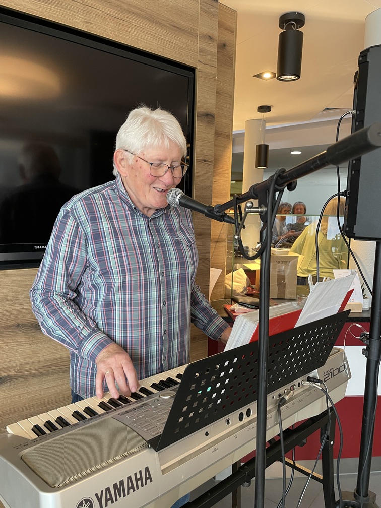 Ein älterer Mann mit weißem Haar und Brille lächelt, während er ein Yamaha-Keyboard spielt und in ein Mikrofon in einem gemütlichen Innenraum singt. Im Hintergrund sind Menschen und Spiegelungen zu sehen.