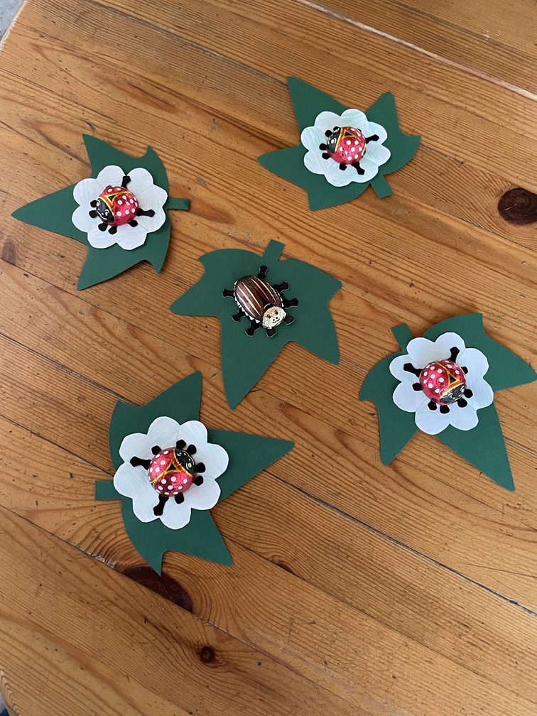 Fünf grüne Papierblätter mit weißen Blüten liegen auf einer Holzfläche. Auf vier Blüten steht jeweils eine rote Marienkäferfigur, auf der fünften eine braune Käferfigur.