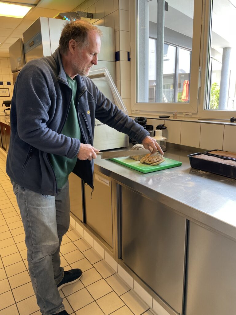 Ein Mann mit Bart schneidet Brot auf einem grünen Schneidebrett in einer modernen Küche mit Edelstahltischen und großen Fenstern.