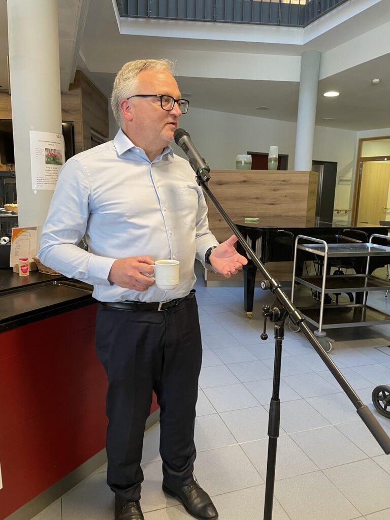 Ein Mann mit Brille und hellblauem Hemd spricht in ein Mikrofon, während er eine weiße Kaffeetasse hält. Er steht in einer modernen Cafeteria mit Tischen, Tabletts und Holzverkleidungen im Hintergrund.