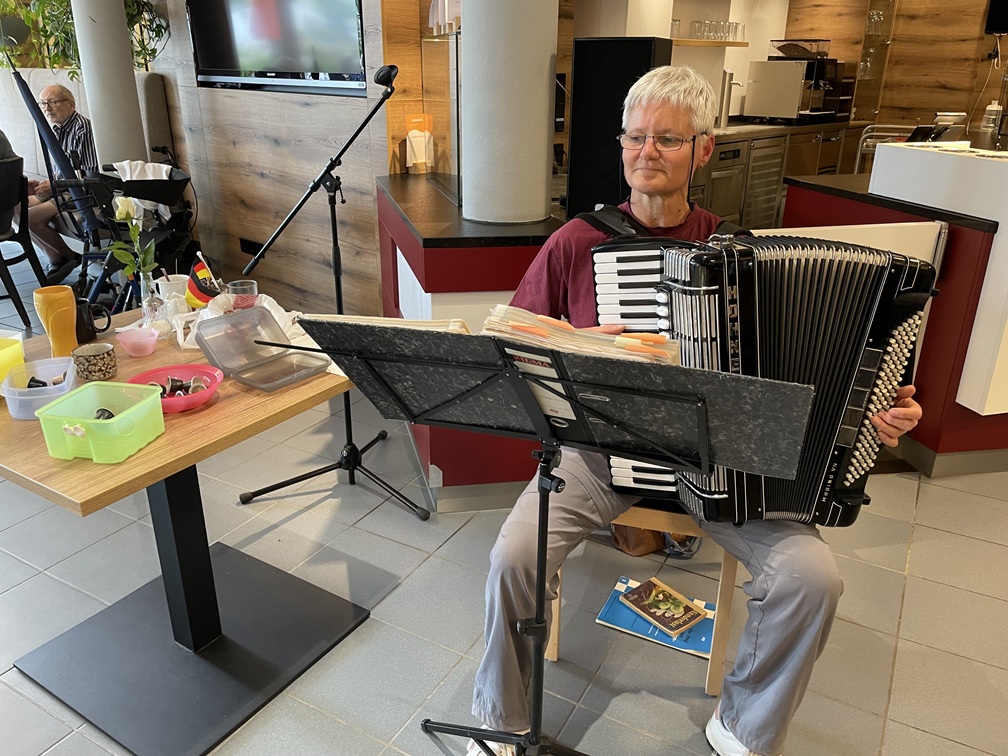 Ein älterer Mann mit grauen Haaren sitzt drinnen und spielt Akkordeon, vor sich liegen Notenblätter. Auf einem Tisch in der Nähe stehen verschiedene Behälter und Getränke, im Hintergrund ist ein Mikrofonständer zu sehen.