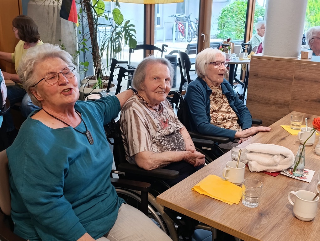 Drei ältere Damen sitzen gemeinsam an einem Tisch in einem hellen Raum, lächeln und genießen die Gesellschaft der anderen. Auf dem Tisch stehen Tassen, Gläser und Servietten, und im Hintergrund sind weitere Personen zu sehen.
