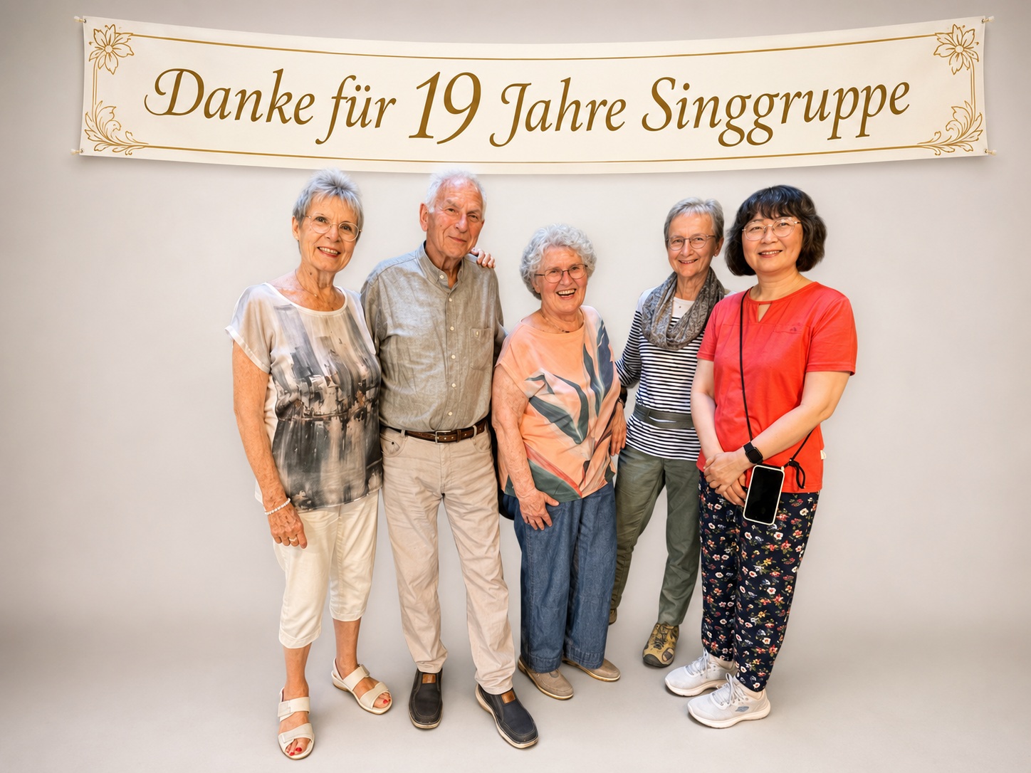 Fünf lächelnde Senioren stehen vor einem Banner mit der Aufschrift „Danke für 19 Jahre Singgruppe“. Sie posieren gemeinsam und wirken glücklich.