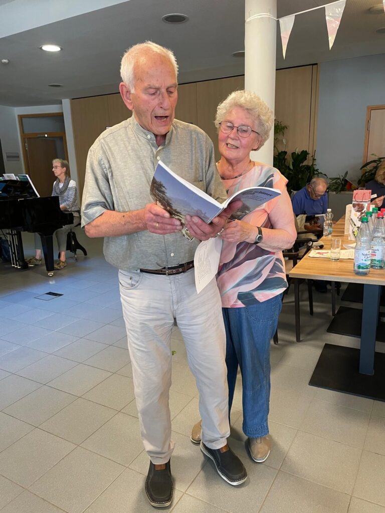 Ein älterer Mann und eine ältere Frau stehen eng beieinander, lächeln und halten ein aufgeschlagenes Buch in den Händen. Es sieht so aus, als würden sie lesen oder singen. Hinter ihnen sitzen Menschen an Tischen, und eine Person spielt Klavier in einem hellen Gemeinschaftsraum.