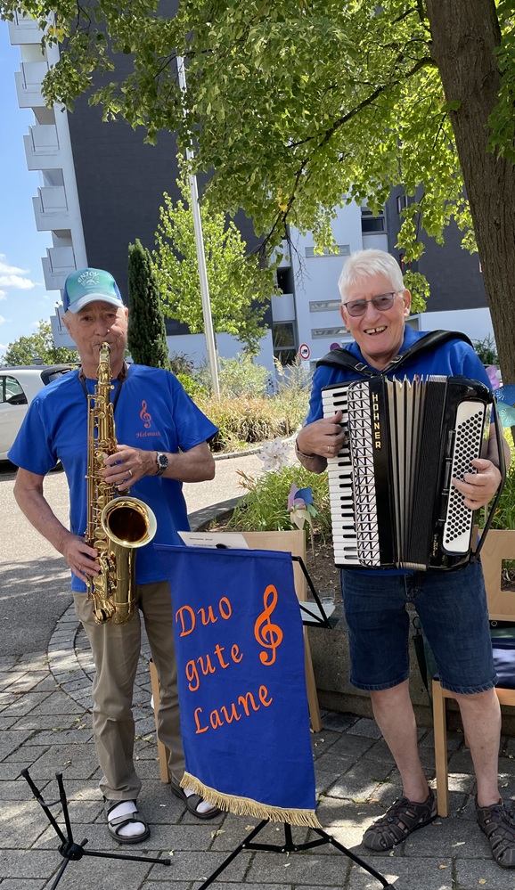 Zwei ältere Männer spielen im Freien Musik; der eine spielt Saxophon, der andere Akkordeon. Sie tragen blaue Hemden und lächeln neben einem Schild mit der Aufschrift „Duo gute Laune“. Im Hintergrund sind Bäume und Gebäude zu sehen.