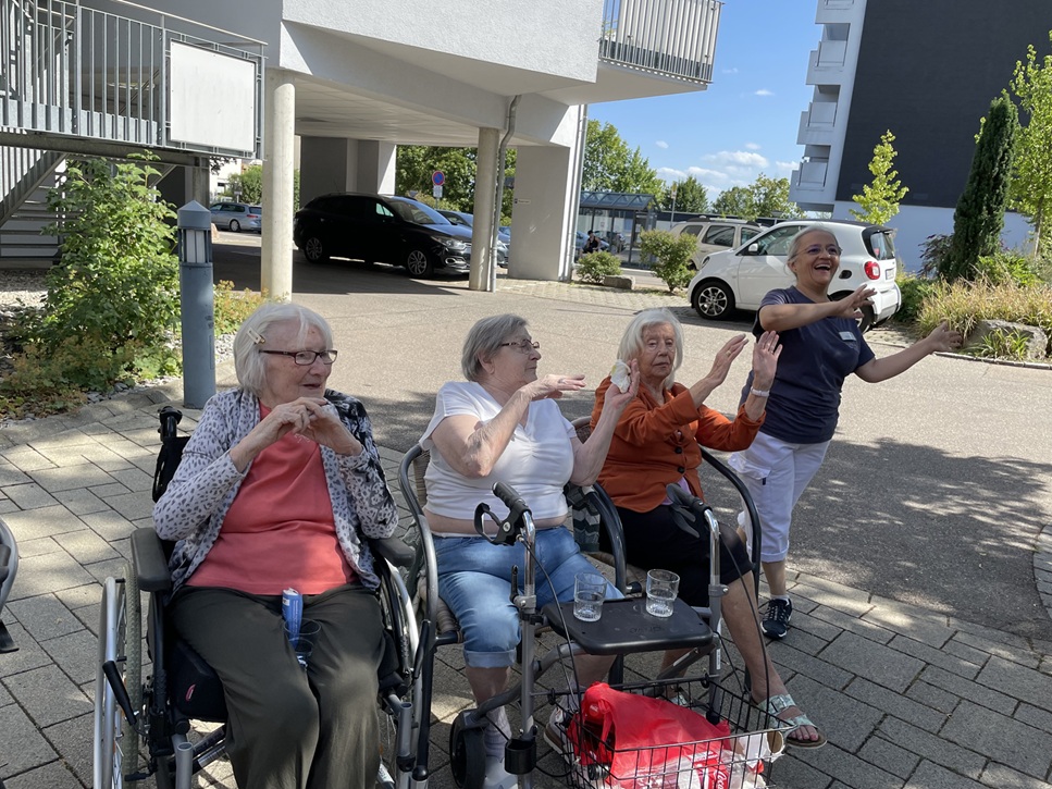 Vier ältere Frauen, zwei sitzen und zwei stehen, befinden sich an einem sonnigen Tag im Freien, lächeln, gestikulieren mit den Händen und genießen die gemeinsame Zeit. Auf dem Bürgersteig stehen neben ihnen Wasserbecher und ein Rollator.