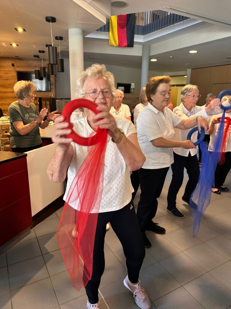 Eine Gruppe älterer Frauen nimmt an einem Indoor-Fitnesskurs teil und hält rote und blaue Ringe mit passenden Schals in den Händen. Sie wirken konzentriert und engagiert, im Hintergrund hängen deutsche Flaggen.
