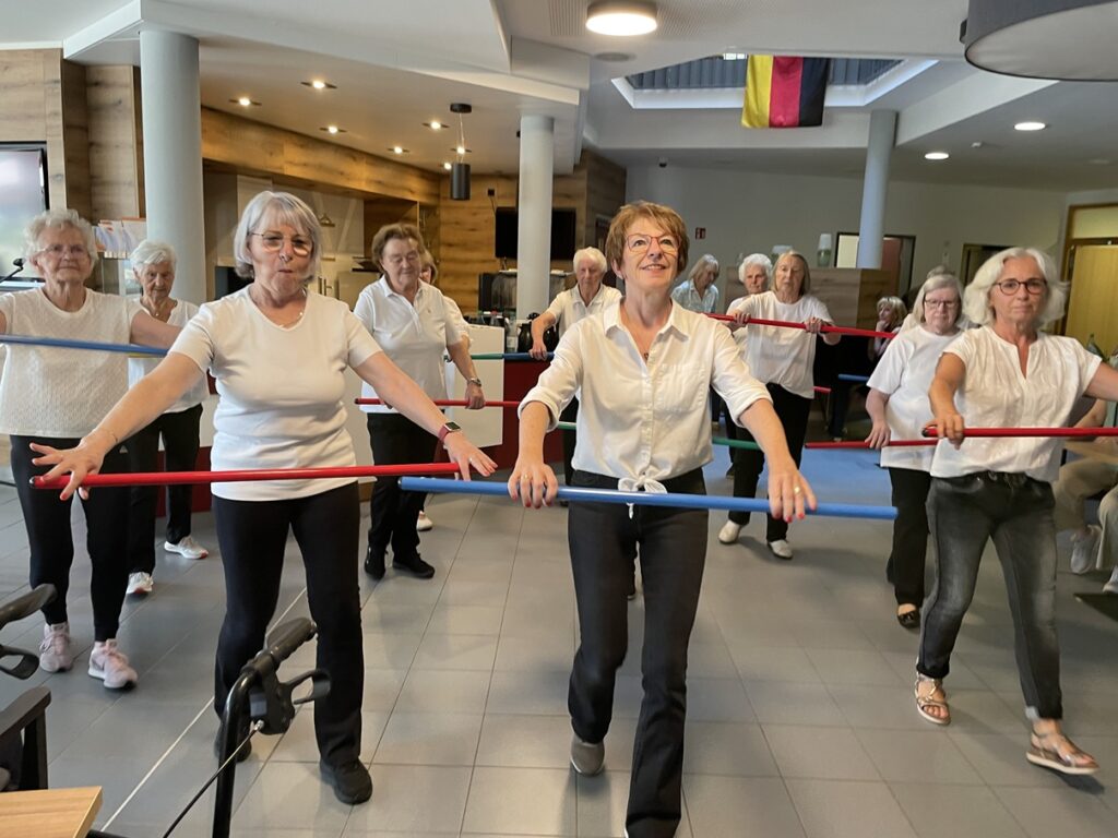 Eine Gruppe älterer Frauen nimmt in einem hellen Raum an einem Fitnesskurs teil. Sie halten lange, bunte Übungsstäbe in den Händen und bewegen sich im Gleichklang. Die meisten tragen weiße Oberteile und dunkle Hosen, und sie scheinen Spaß an der Aktivität zu haben.