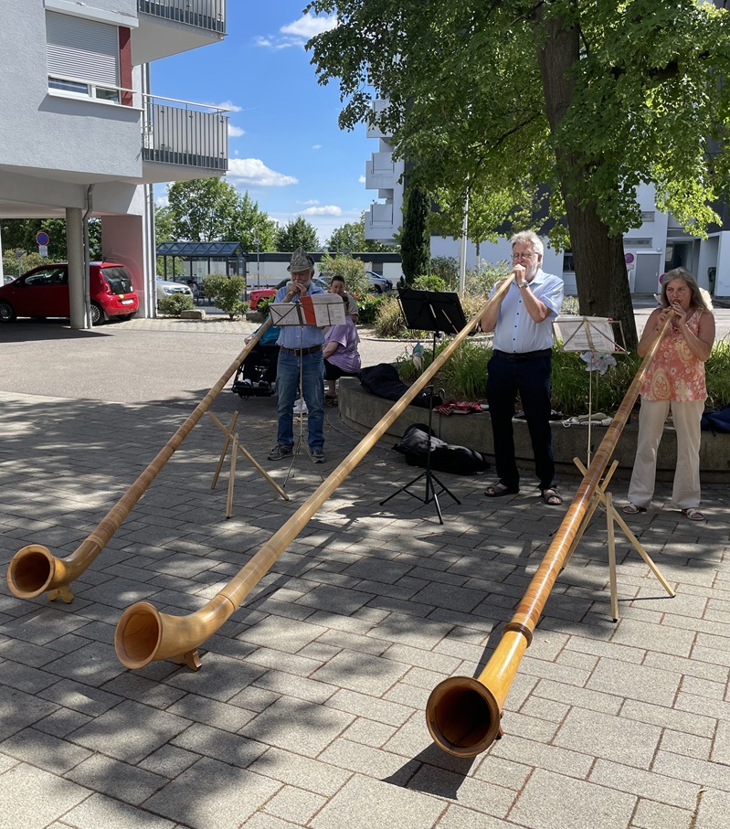 Drei Menschen stehen im Freien und spielen auf langen hölzernen Alphörnern. Sie stehen neben Notenständern auf einer gepflasterten Fläche in der Nähe von Bäumen und Gebäuden unter einem strahlenden, teilweise bewölkten Himmel.