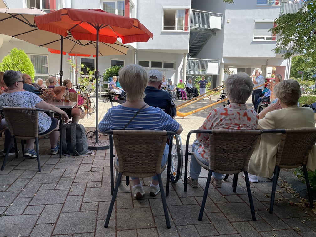Eine Gruppe älterer Menschen sitzt an einem sonnigen Tag im Freien unter roten Sonnenschirmen und beobachtet Musiker, die im Innenhof eines Wohnhauses auf langen hölzernen Alphörnern spielen.
