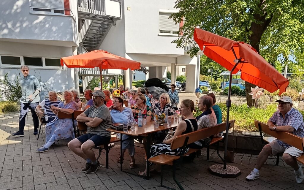 Eine Gruppe von Menschen sitzt draußen an Tischen unter orangefarbenen Sonnenschirmen und genießt einen sonnigen Tag in der Nähe eines weißen Gebäudes, im Hintergrund sind Bäume und Grünflächen zu sehen.