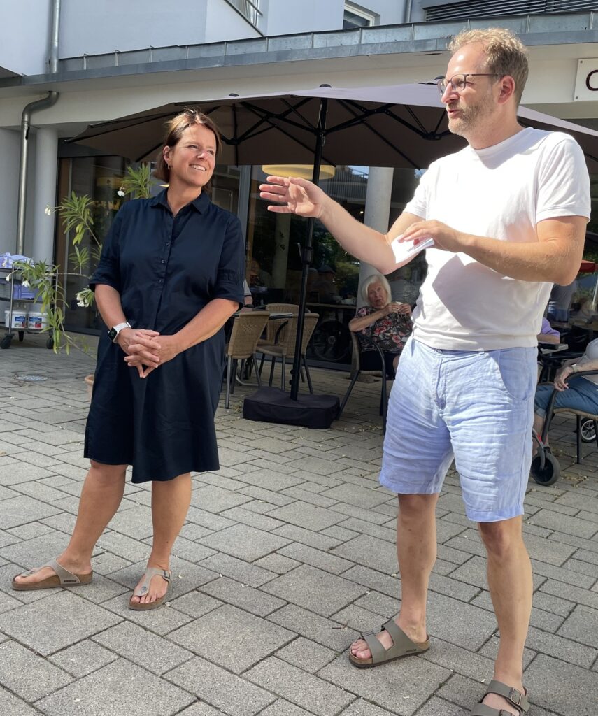 Eine Frau in einem schwarzen Kleid und ein Mann in einem weißen Hemd und blauen Shorts stehen draußen auf einer Terrasse. Der Mann gestikuliert beim Sprechen, und die Frau lächelt. Im Hintergrund sitzen Menschen.