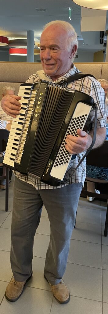 Ein älterer Mann in einem karierten Hemd und einer grauen Hose lächelt, während er in einem Restaurant auf einem schwarz-weißen Hohner-Akkordeon spielt.