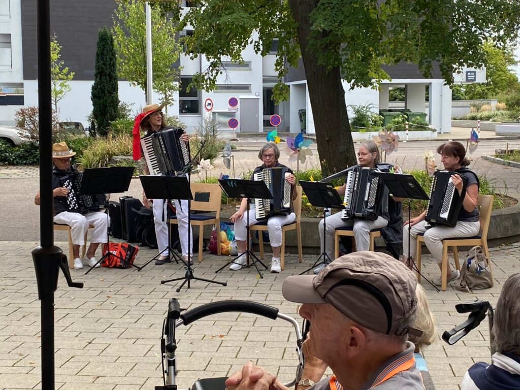 Fünf Personen sitzen im Freien und spielen in einer kleinen Gruppe Akkordeon. Vier von ihnen sind weiß gekleidet, eine steht mit Hut und rotem Schal da. Im Vordergrund schauen zwei ältere Menschen zu, von denen einer einen Rollator benutzt. Im Hintergrund sind Bäume und Gebäude zu sehen.