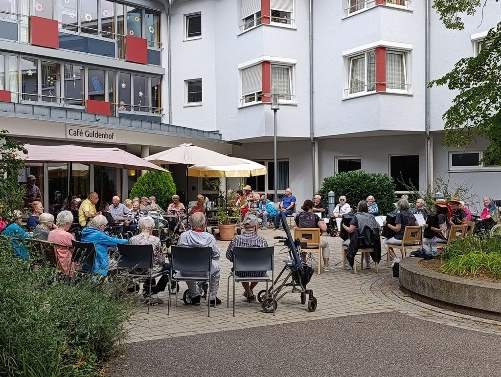Eine Gruppe älterer Menschen sitzt auf Stühlen, unterhält sich und entspannt sich im Innenhof vor einem Gebäude mit der Aufschrift „Café Guldenhof“. Einige benutzen Rollatoren, und rundherum stehen Sonnenschirme und Pflanzen.