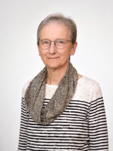 Eine ältere Frau mit kurzen grauen Haaren, Brille und ausdruckslosem Gesicht trägt ein gestreiftes Hemd und einen gemusterten Schal und posiert vor einem schlichten hellen Hintergrund.