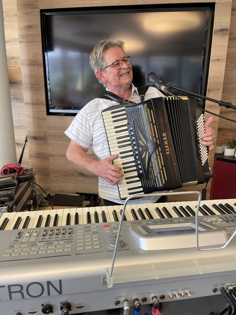 Ein Mann mit Brille und gestreiftem Hemd steht in einem modernen Raum hinter einem Keyboard, spielt Akkordeon, singt ins Mikrofon und lächelt dabei.