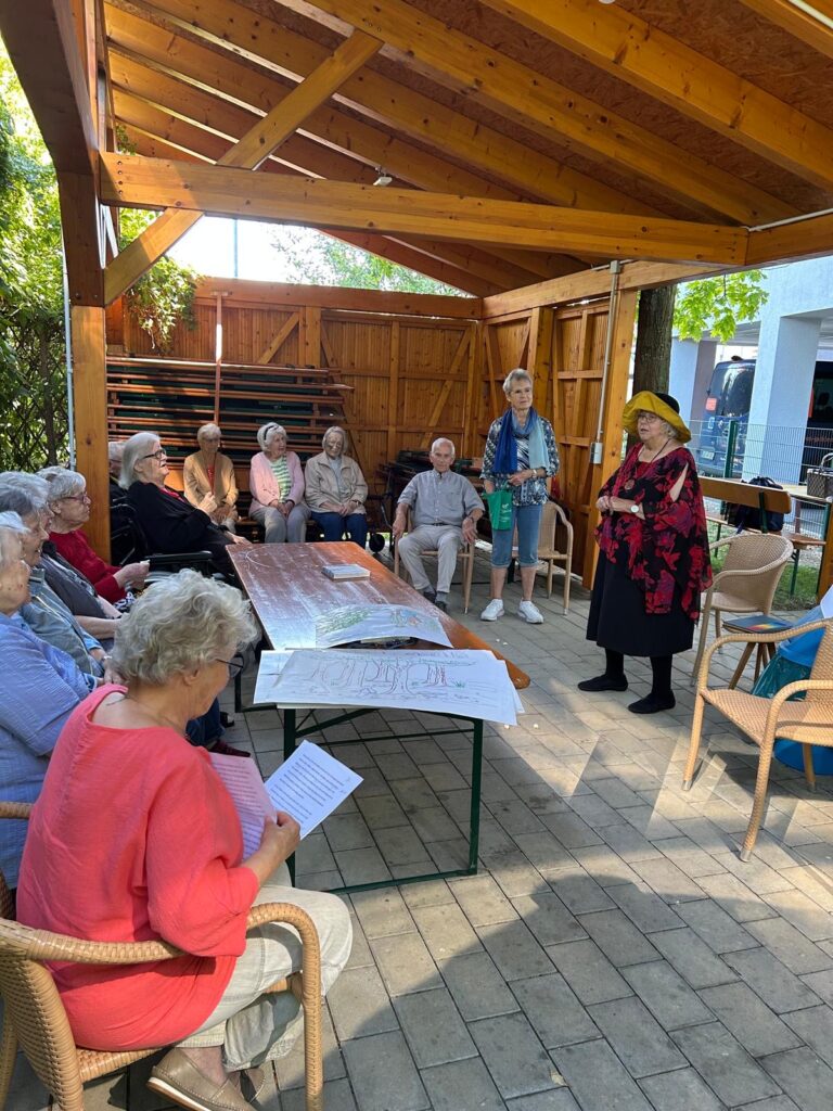 Eine Gruppe älterer Menschen sitzt auf Stühlen unter einem Holzpavillon und hört einer Frau mit Hut und rotem Schal zu. Auf dem Tisch in der Mitte liegen Papiere und eine große Karte. Sonnenlicht fällt durch die offenen Seiten herein.