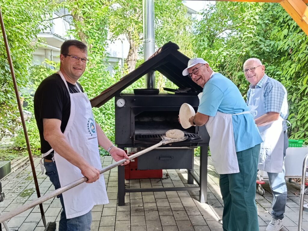 Drei Männer in Schürzen lächeln, während sie draußen Brot backen – in einem Holzofen, der von Grünpflanzen und Ziegelsteinen umgeben ist. Einer hält den Teig in der Hand, ein anderer bedient die Backschaufel, und der dritte steht daneben; alle wirken fröhlich und konzentriert.