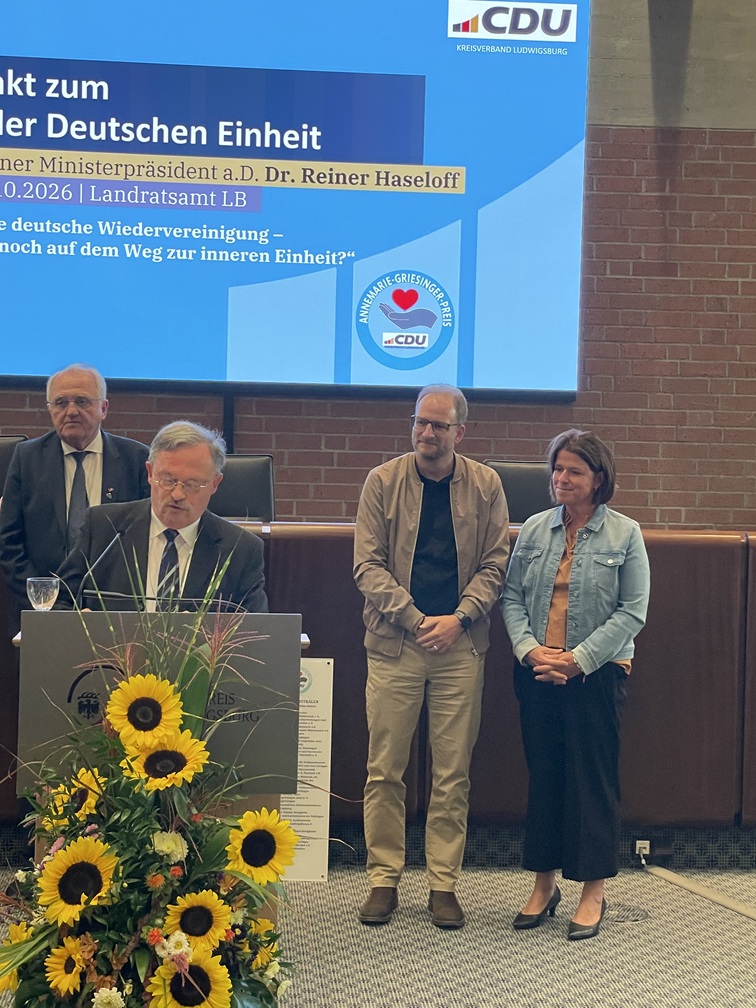 Ein Mann steht auf einem Podium und hält vor einer Leinwand mit dem CDU-Logo eine Rede, während neben ihm ein Mann und eine Frau stehen.