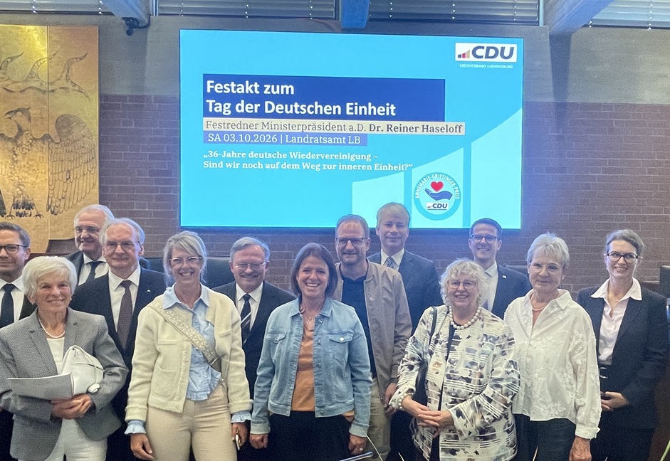 Eine Gruppe von Menschen steht vor einem digitalen Bildschirm, auf dem Informationen zu einer Feier zum Tag der Deutschen Einheit mit dem Redner Dr. Reiner Haseloff angezeigt werden.