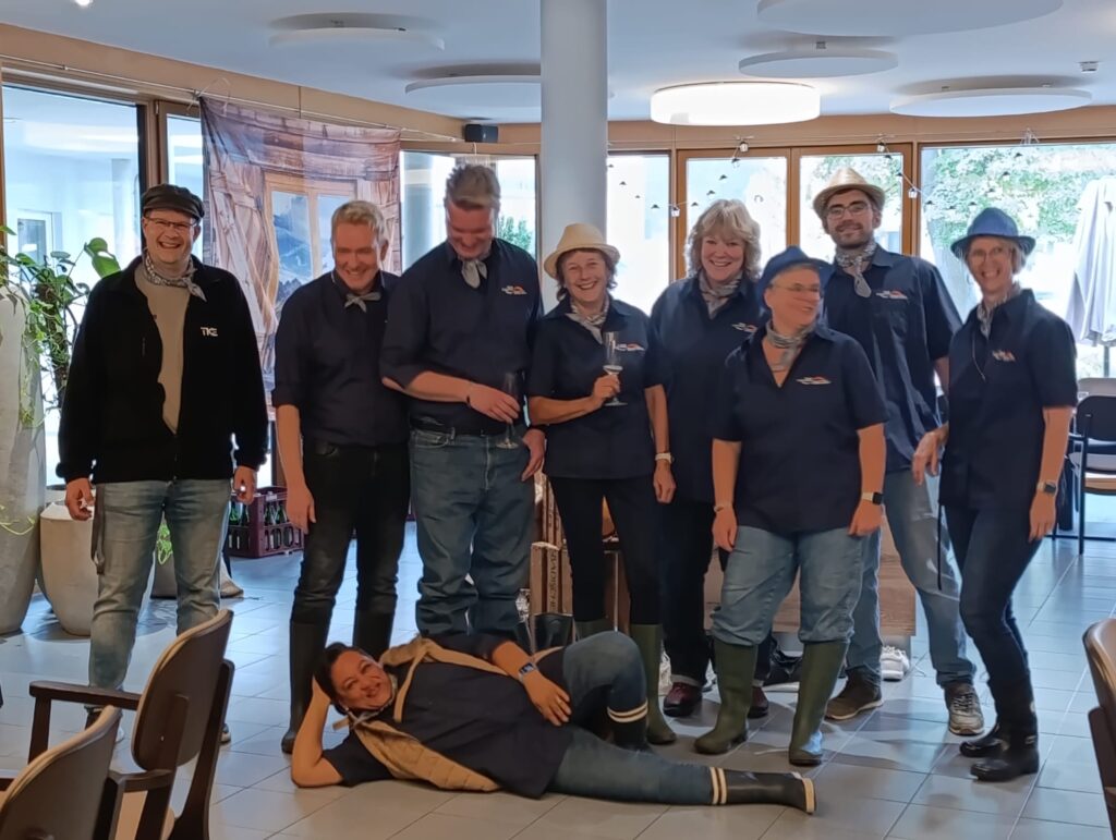 Eine Gruppe von neun Personen, die alle einheitliche marineblaue Hemden und Halstücher tragen, posiert gemeinsam in einem Raum, wobei sich im Vordergrund eine Person auf dem Boden ausstreckt.