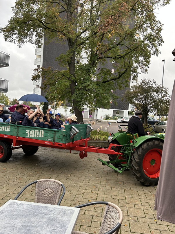 Ein grüner Traktor zieht einen grünen Anhänger, auf dem eine Gruppe von Menschen steht und winkt; einer von ihnen hält einen blauen Regenschirm in der Hand.