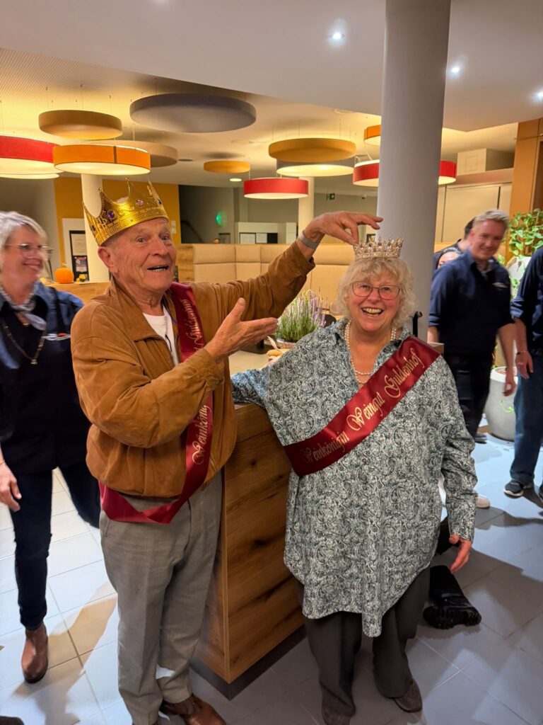 Ein älterer Mann mit einer goldenen Krone setzt einer lächelnden älteren Frau, die eine rote Schärpe mit der Aufschrift Weingut Guldenhof trägt, eine kleinere Krone auf den Kopf.