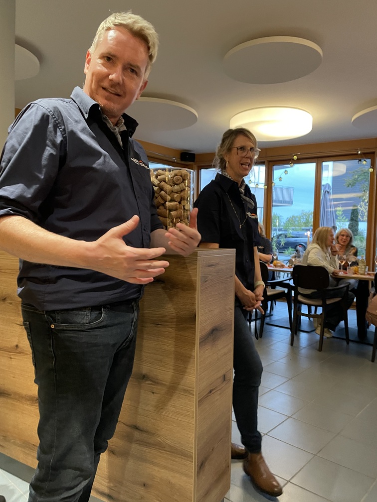 Ein Mann, der in der Nähe einer mit Weinkorken gefüllten Theke gestikuliert, während eine Frau daneben steht und im Hintergrund Gäste an Tischen sitzen.