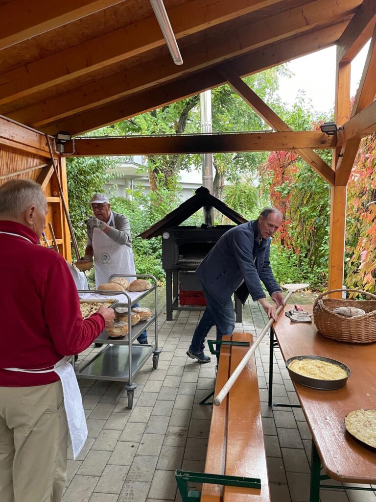 Drei Männer arbeiten gemeinsam unter einem Holzpavillon daran, Brot in einem Außenofen zu backen.