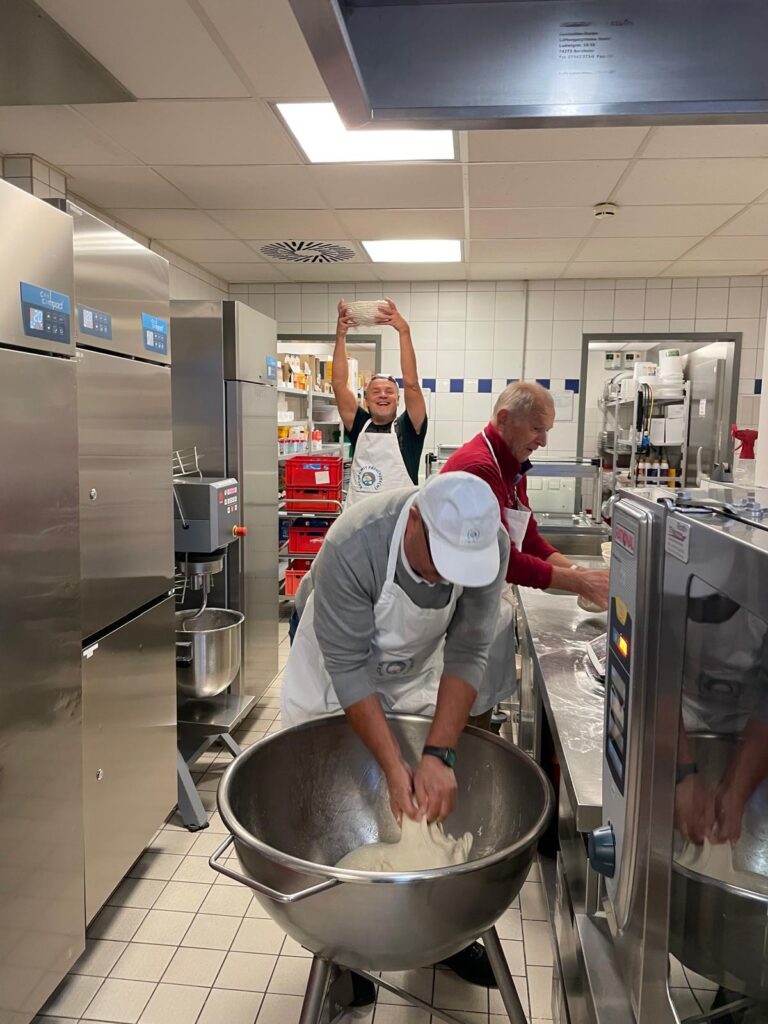 Drei Bäcker in Schürzen arbeiten in einer Großküche; einer von ihnen hält einen Teigball über den Kopf.